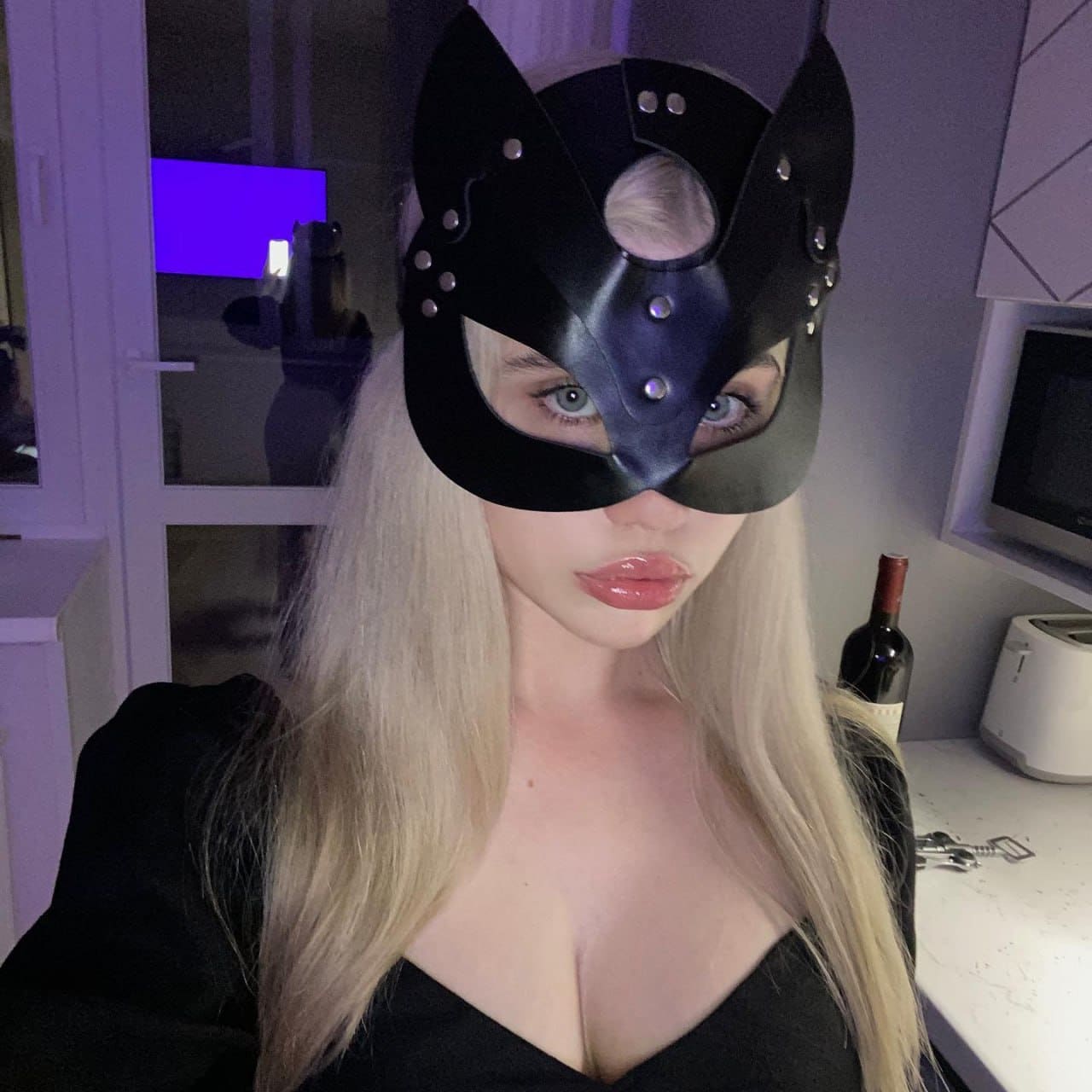Девушка в маске кота с шипами и черном топе селфирует на кухне, на фоне вино и тостер.